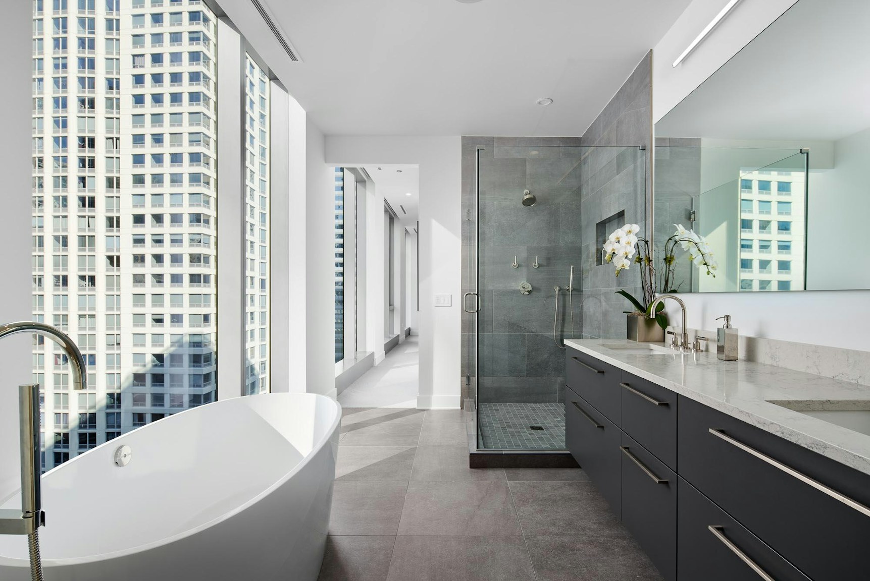 A modern bathroom with a freestanding white tub, floor-to-ceiling windows offering city views, gray tile floors, and a glass-enclosed shower with a sleek design.